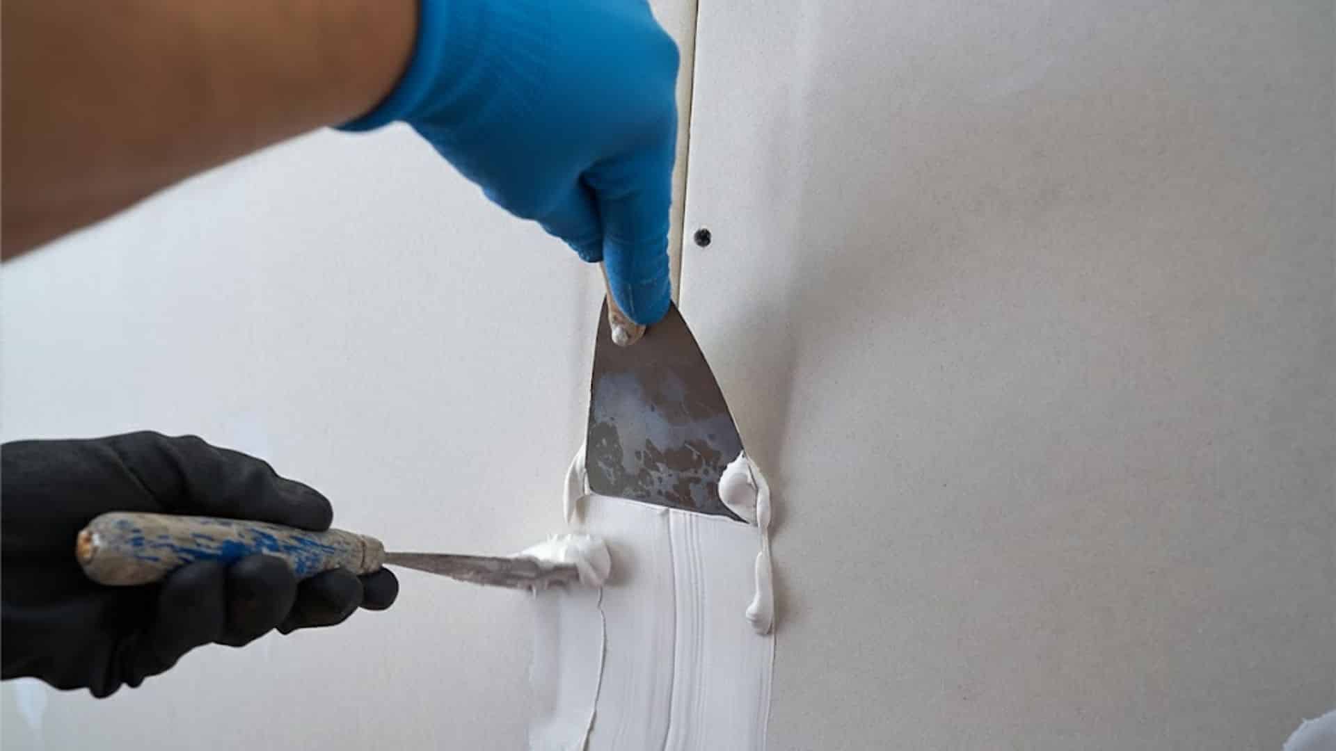 Hands in blue and black gloves use putty knives to apply smooth white plaster over a drywall seam