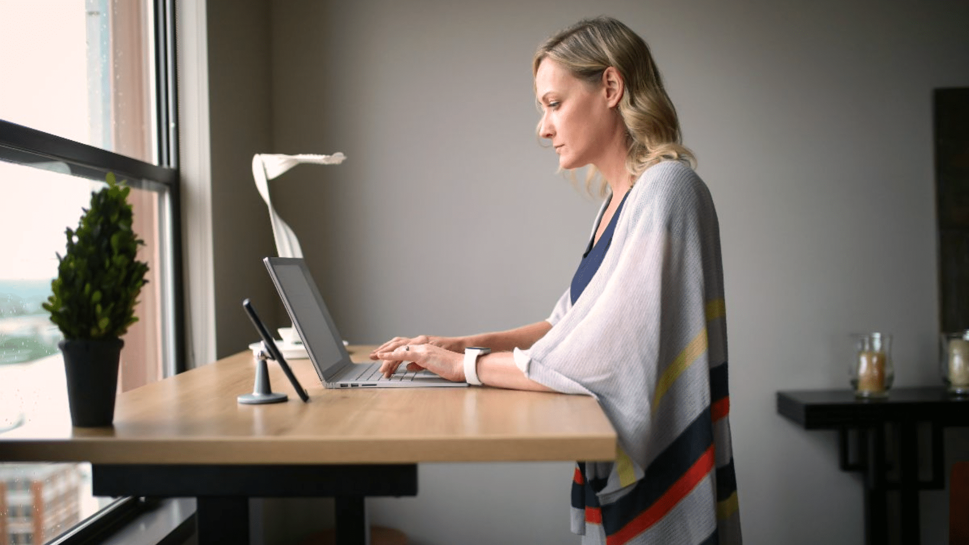woman typing on a laptop at a wooden standing desk beside a large window