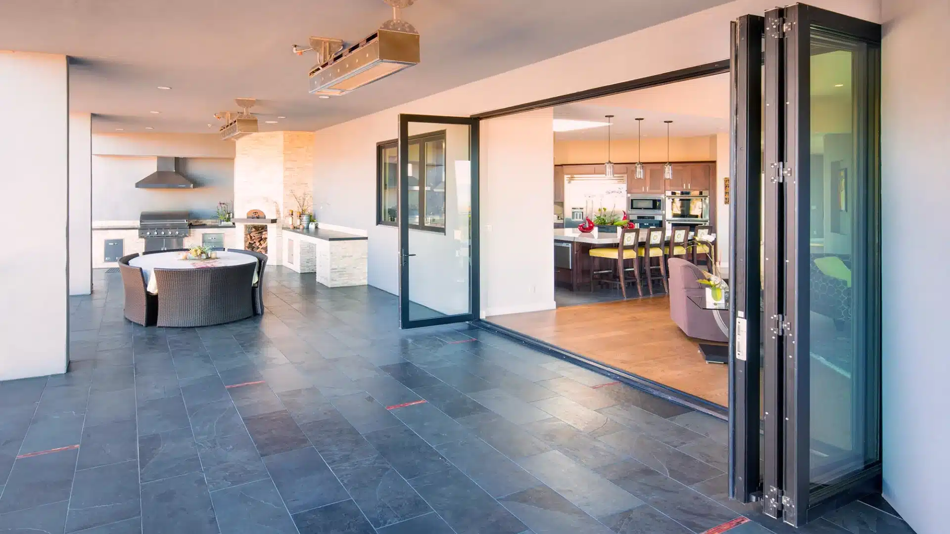Bi-fold glass doors open to a modern patio with outdoor kitchen, dining area, and seamless indoor connection