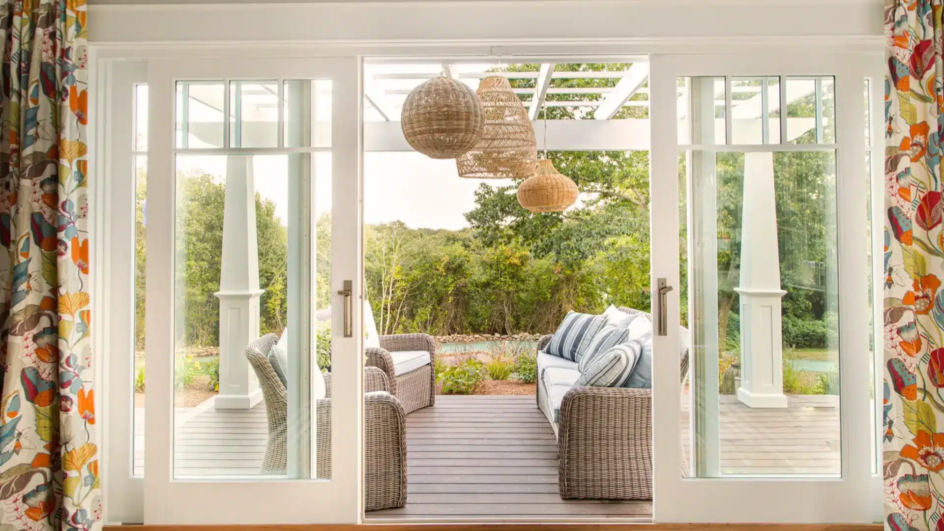 Sliding glass doors open to a cozy patio with wicker seating, hanging lights, and a garden view beyond