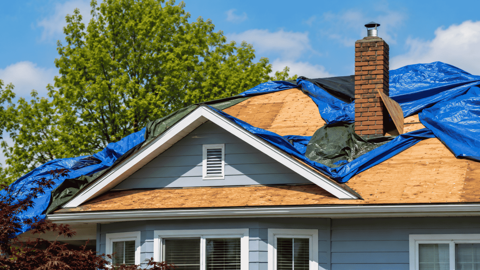 Storm-damaged suburban home with roof tarp, missing shingles, fallen branches, and debris after severe weather