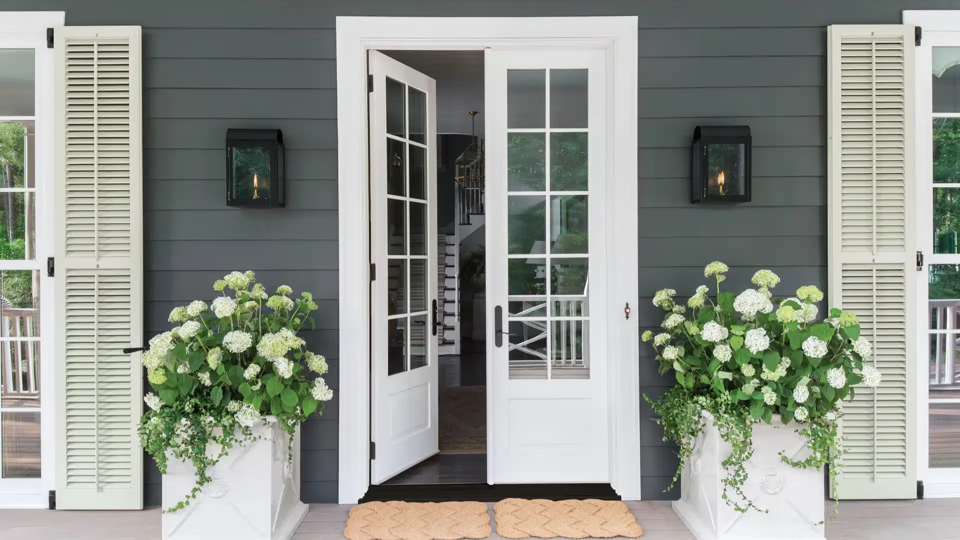 White French doors open to a gray house exterior with shutters, wall lanterns, and white hydrangea planters on porch
