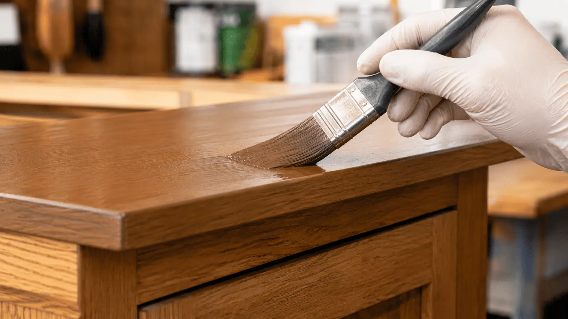 Gloved hand applying brown paint to the top of a wooden cabinet.