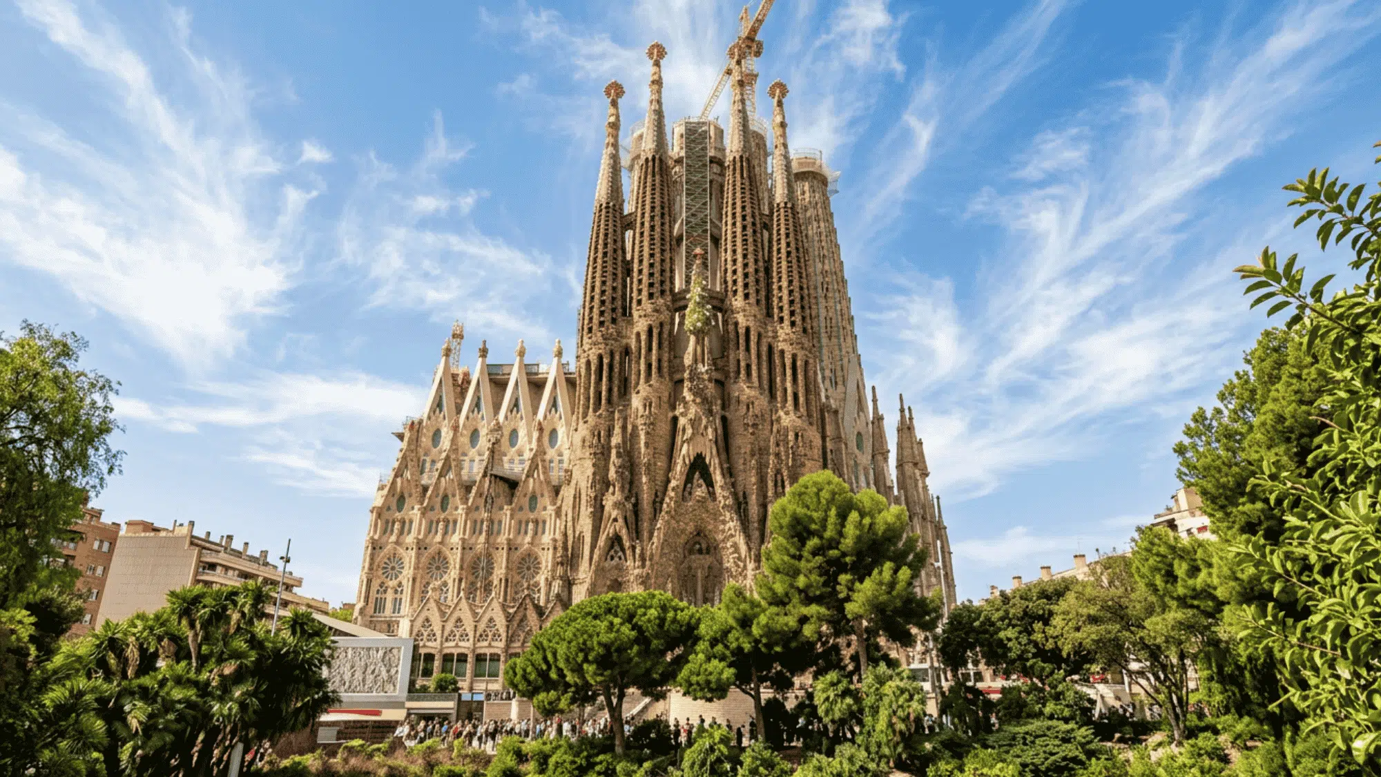 Sagrada Família in Barcelona with soaring spires, stone façades, lush gardens, and a bright blue sky.